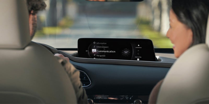 Two people inside a car, with the infotainment screen showing 'Communication' highlighted, and 'Apple CarPlay' visible.
