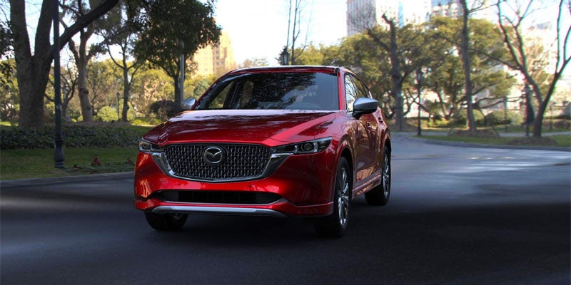 A shiny red Mazda CX-5 SUV parked on a paved road, surrounded by trees and distant buildings.