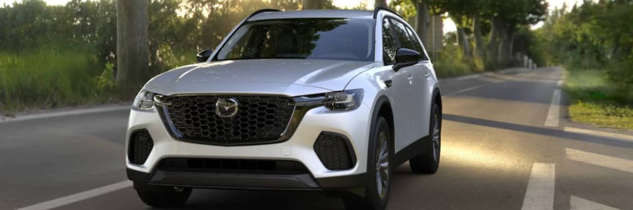 Silver Mazda CX-90 SUV parked on a tree-lined road, with sunlight reflecting on the asphalt.