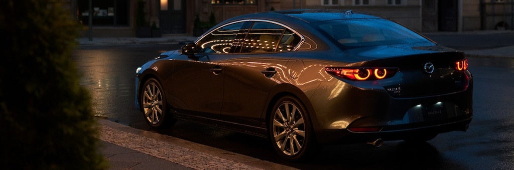 Dark gray Mazda parked on a wet city street at night, with reflections of lights shimmering on its body.