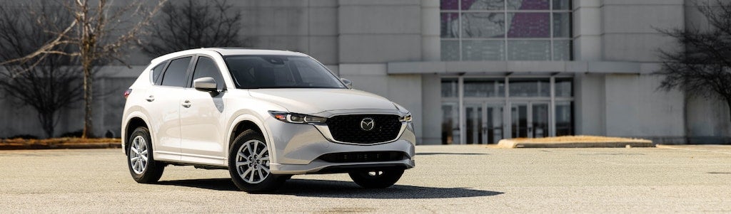 White Mazda CX-5 SUV parked in a lot in front of a gray building with glass doors and bare trees.