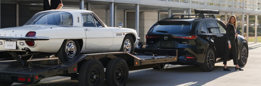 White vintage Mazda Cosmo on a flatbed trailer towed by a black modern Mazda SUV with a woman standing beside it.