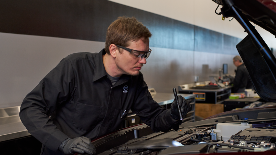 A Mazda service tech leaning over a car with its hood opened, shining a flashlight on the engine